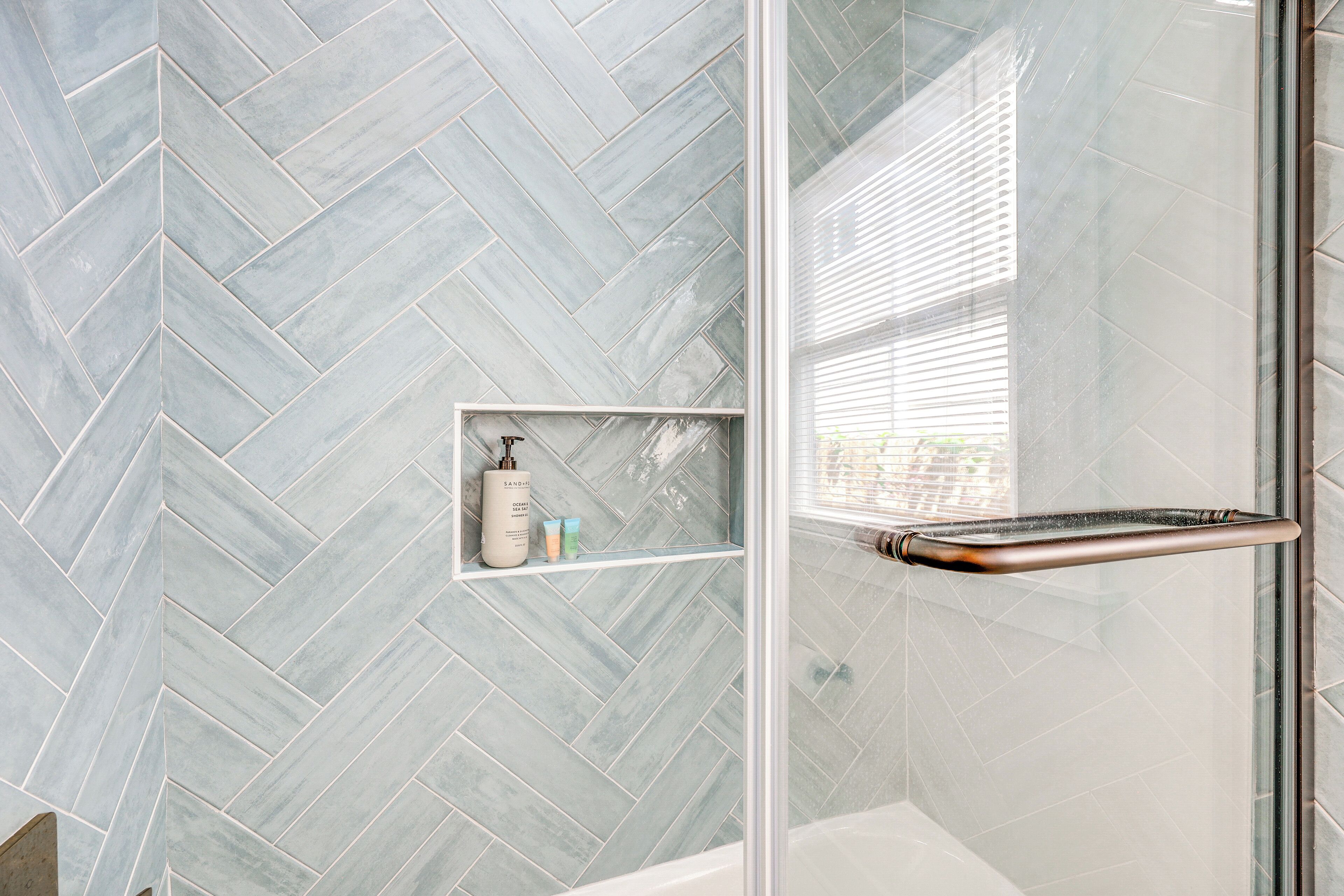 A bathroom with a herringbone tile pattern, a glass shower enclosure, and a shelf with toiletries.