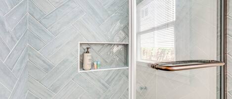 A bathroom with a herringbone tile pattern, a glass shower enclosure, and a shelf with toiletries.