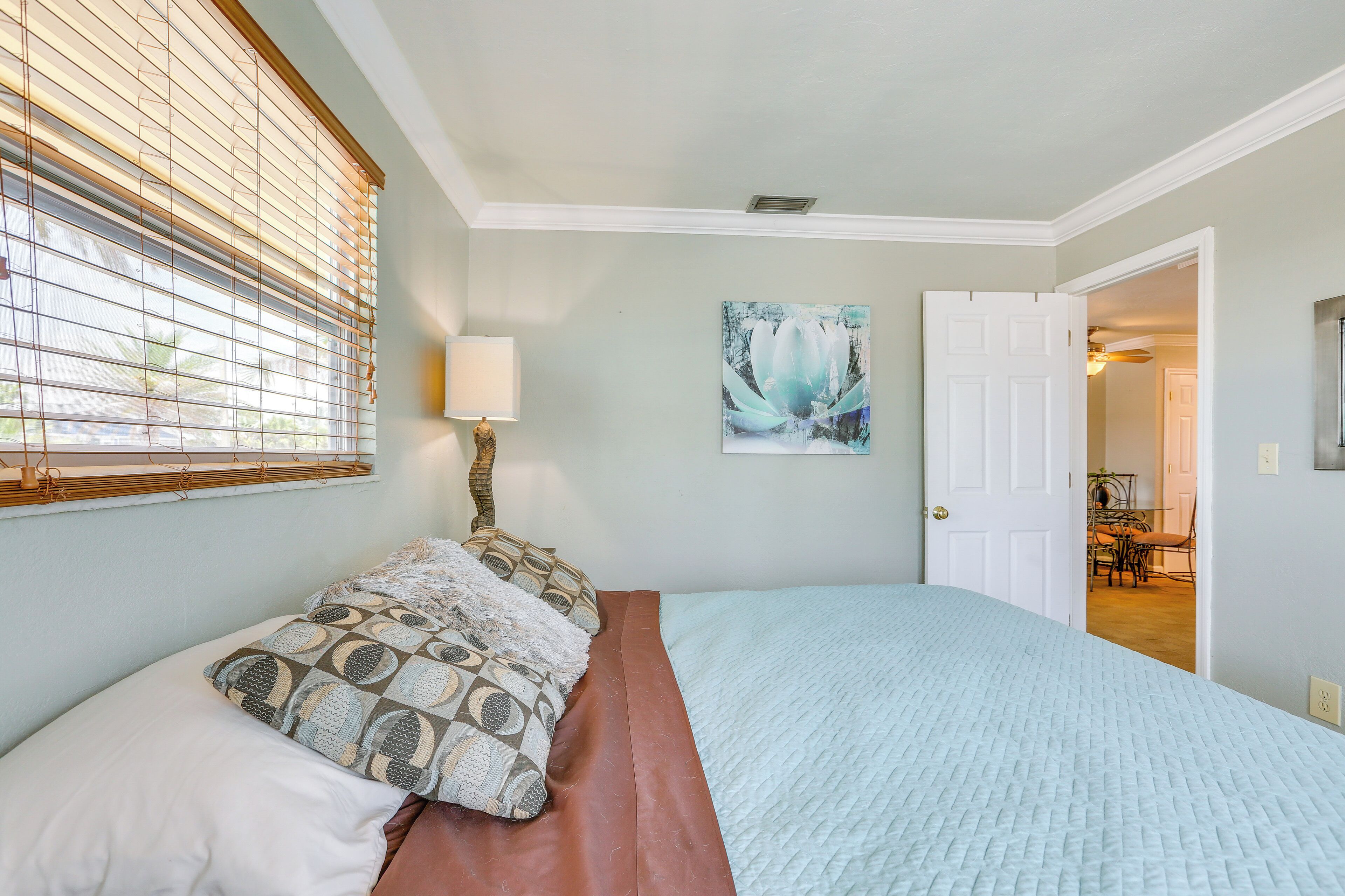 A bedroom with a bed, a window with blinds, a painting on the wall, and a door leading to another room.