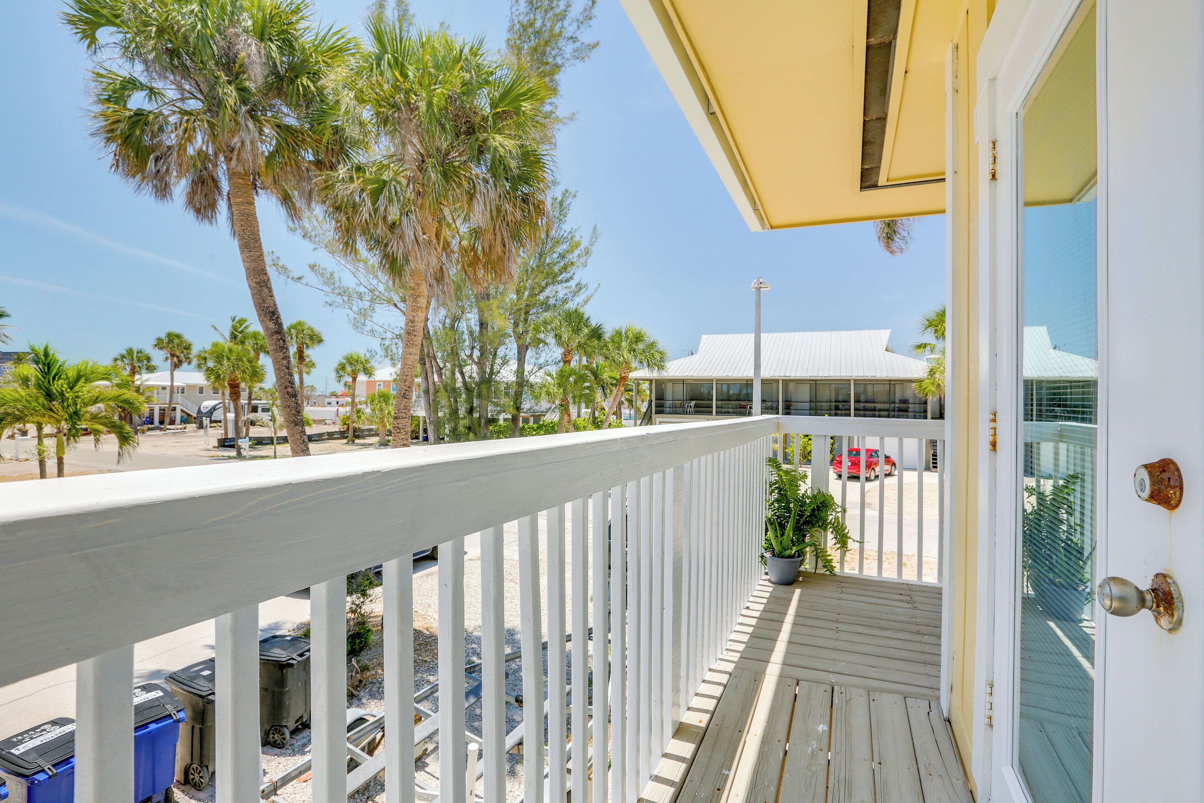 A balcony with a white railing, overlooking a street with palm trees and a building with a white roof.