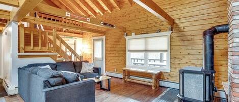A cozy cabin interior with a wooden ceiling, a sofa, a bench, and a wood-burning stove.