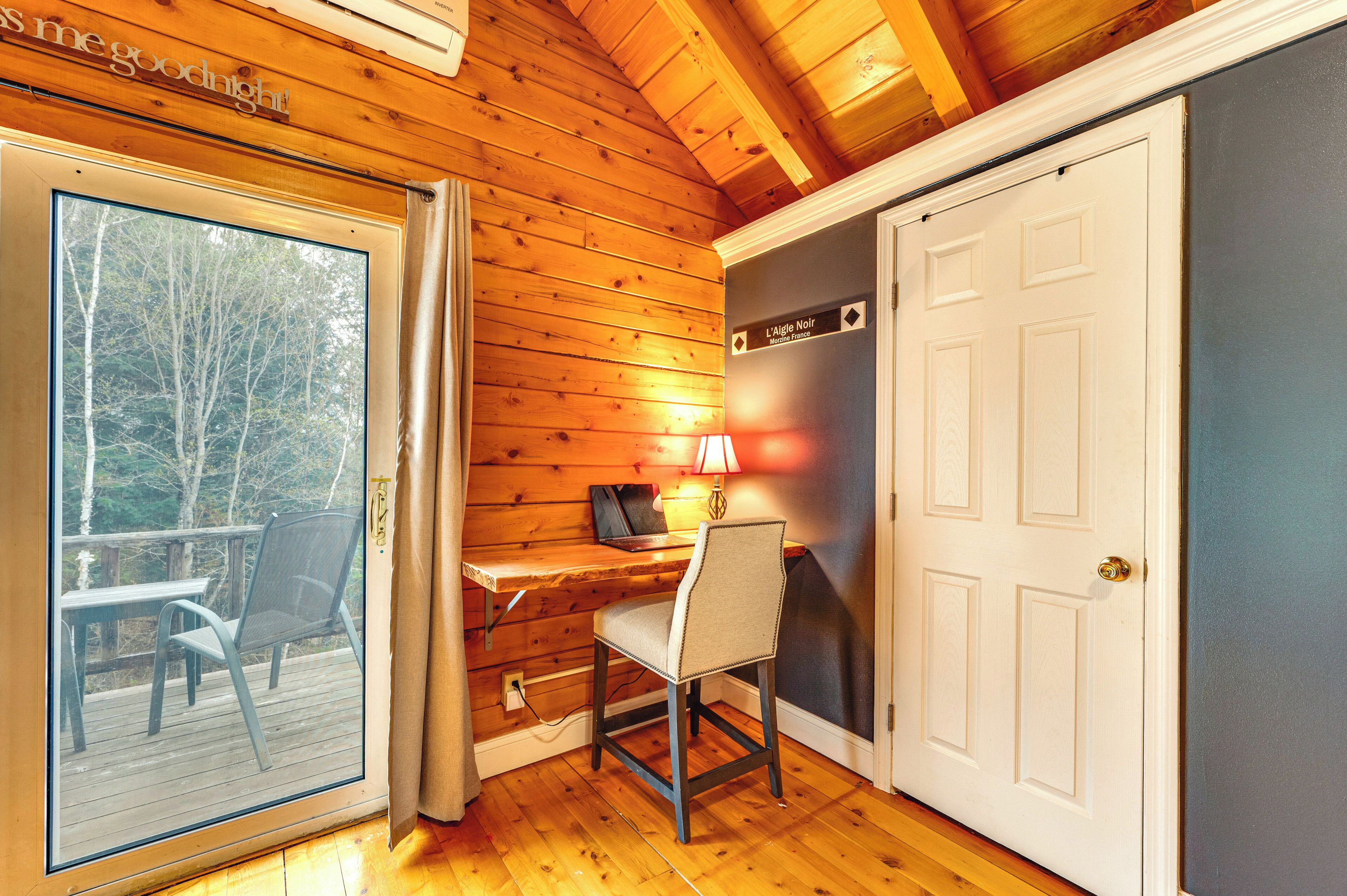 A room with a wooden interior, a desk with a chair, a door, and a window overlooking a deck.