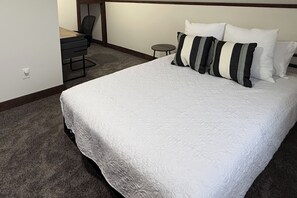 Een net opgemaakt bed met een witte quilt en gestreepte kussens, een klein rond tafeltje, en een bureau met een stoel in een hotelkamer.