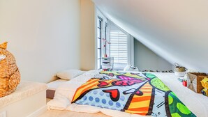 Une chambre avec un plafond en pente, un lit recouvert d’une couette aux couleurs vives, un panier en osier et une fenêtre avec des stores.