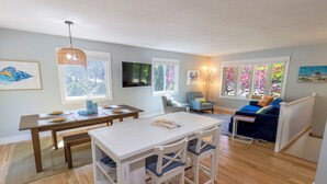 Un salón moderno con zona de comedor, suelo de madera y ventanas amplias con flores rosadas.