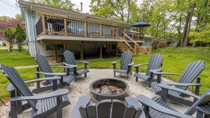 Una casa con terraza, mobiliario exterior y sillas tipo Adirondack, además de una fogata.