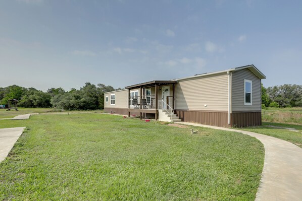 A single-story mobile home with a front porch and a small deck, surrounded by a grassy yard and a concrete path.