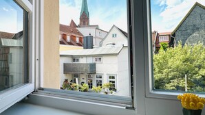 Blick aus einem Fenster auf eine Kirche mit Kirchturm, angrenzende Gebäude und einen Balkon mit Blumentöpfen.