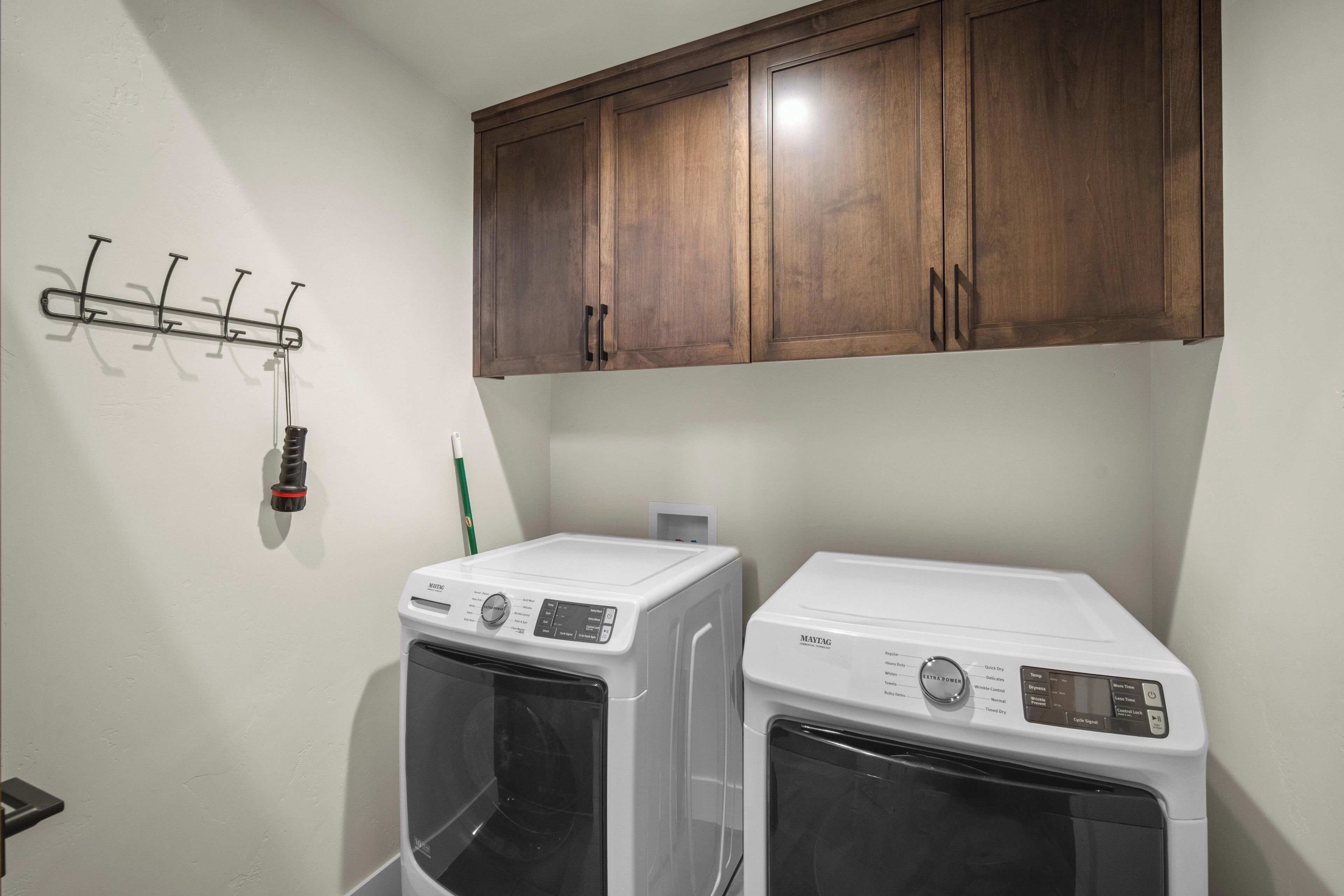 A laundry room with two stacked front-loading washing machines, a wooden cabinet above, and a metal drying rack with hooks.