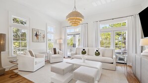 Amplia sala de estar con un sofá blanco, dos sillones y una mesa de centro. Cuenta con ventanas grandes con cortinas blancas, una lámpara colgante y vistas al exterior con vegetación.