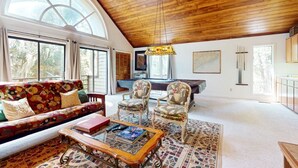 Amplia sala de estar con techo de madera alto, ventanas grandes, un sofá colorido, dos sillones, una mesa de centro y una mesa de billar.