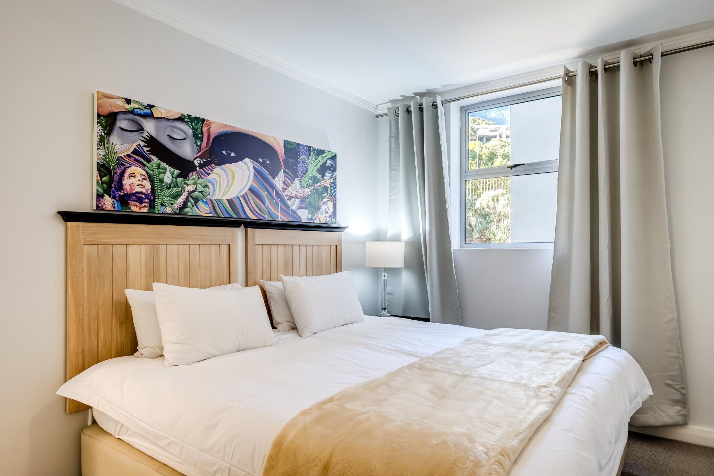 Una habitación de hotel con una cama grande, un cabecero de madera, un cuadro llamativo sobre la cama, una ventana con cortinas y una lámpara de noche.