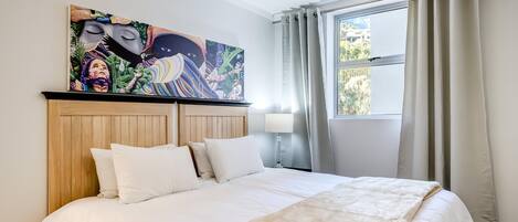 Una habitación de hotel con una cama grande, un cabecero de madera, un cuadro llamativo sobre la cama, una ventana con cortinas y una lámpara de noche.
