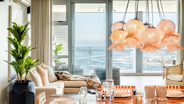 Una sala de estar moderna con un amplio ventanal que da a un paisaje urbano, un comedor con muebles de madera y una planta en maceta.