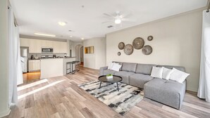Ein modernes Wohnzimmer mit einem grauen Sofa, einem Couchtisch und einem gemusterten Teppich. Im Hintergrund ist eine Küchenzeile mit weißen Schränken und einer Mikrowelle zu sehen.