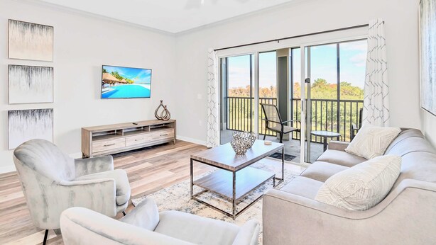 A modern living room with a sofa, armchairs, a coffee table, and a TV. There is a balcony with a view of trees and a railing. - Modern 2BR Condo w Private Patio7 min to Disney (Winter Garden)