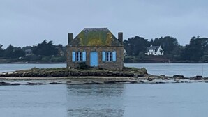 Une maison en pierre avec une porte et des fenêtres bleues, située sur un promontoire rocheux au bord d’une étendue d’eau.