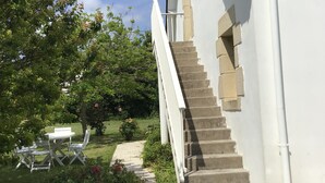 Un bâtiment blanc avec un escalier en pierre menant à un deuxième étage, entouré de verdure et doté d’un petit espace extérieur aménagé pour s’asseoir.