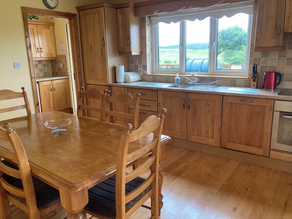 A kitchen with wooden cabinets, a dining table with chairs, a window with a view of the countryside, and a red kettle on the counter. - Burkes Holiday Cottages (Rathnamagh)