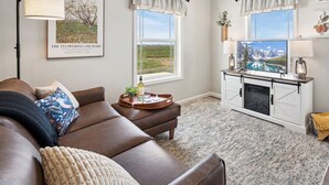 A living room with a brown leather couch, a small table with a bottle and glasses, a TV on a cabinet, and a view of mountains through the window.