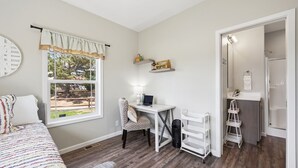 A bedroom with a bed, a desk with a laptop, a chair, a window with curtains, and a bathroom visible through an open door.