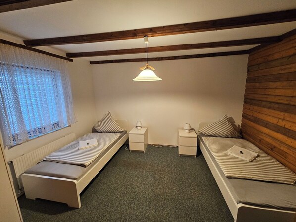 A small, cozy room with two single beds, a window with lace curtains, and a wooden ceiling.