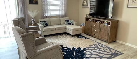 A living room with a sectional sofa, a small ottoman, a wooden entertainment center with a TV, and a decorative rug.