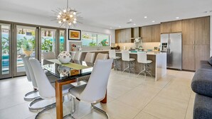 A modern kitchen with a central island, bar stools, and a dining area with a glass table and chairs. - The Cove: Heated Pool, BBQ, Walk To Downtown PS (Palm Springs)