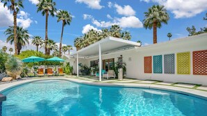 A swimming pool with a covered patio area, palm trees, and a wall with decorative panels. - The Cove: Heated Pool, BBQ, Walk To Downtown PS (Palm Springs)