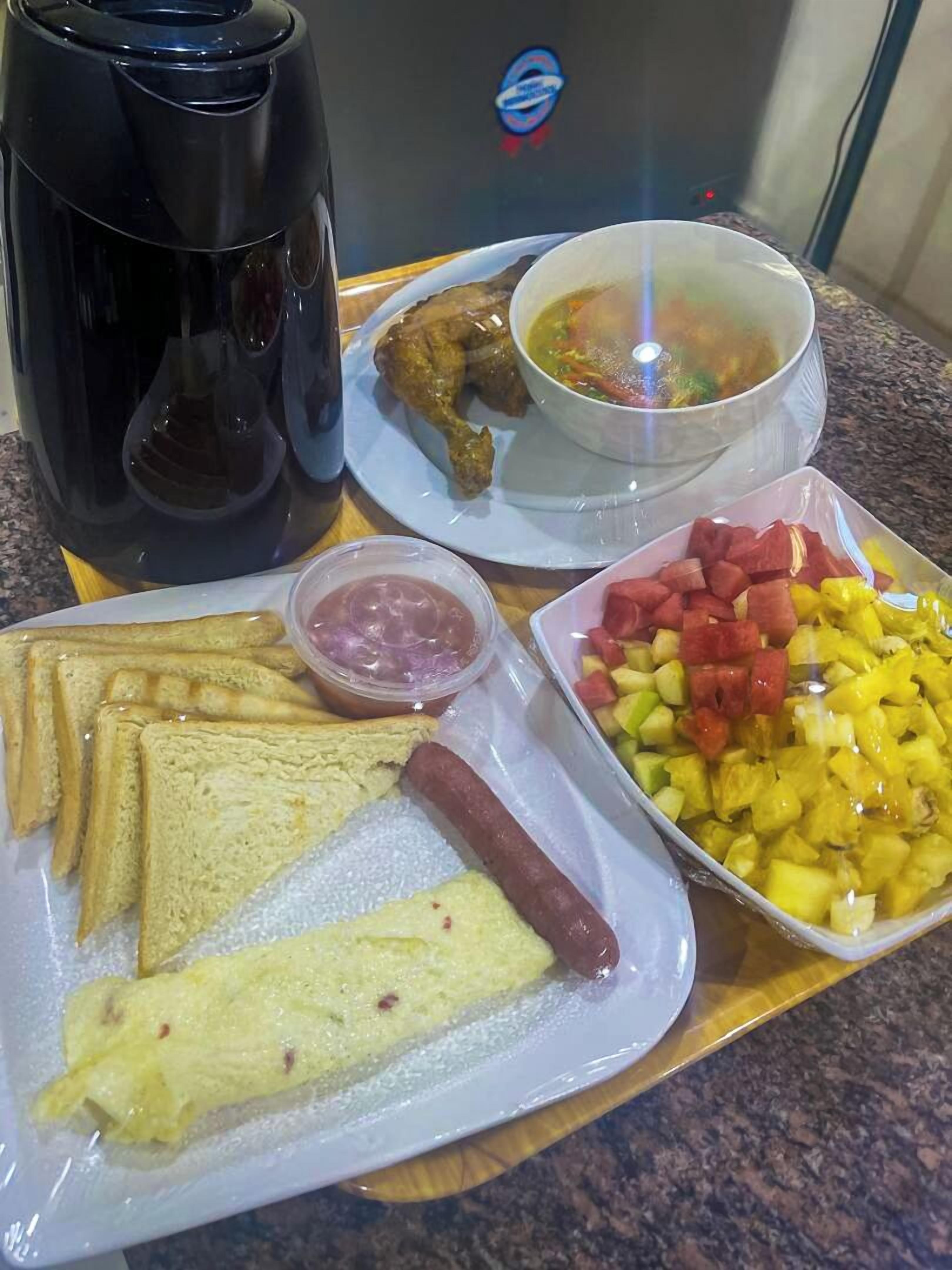 A tray with a hot dog, sliced bread, a small container of ketchup, a bowl of soup, and a dish of diced fruit.