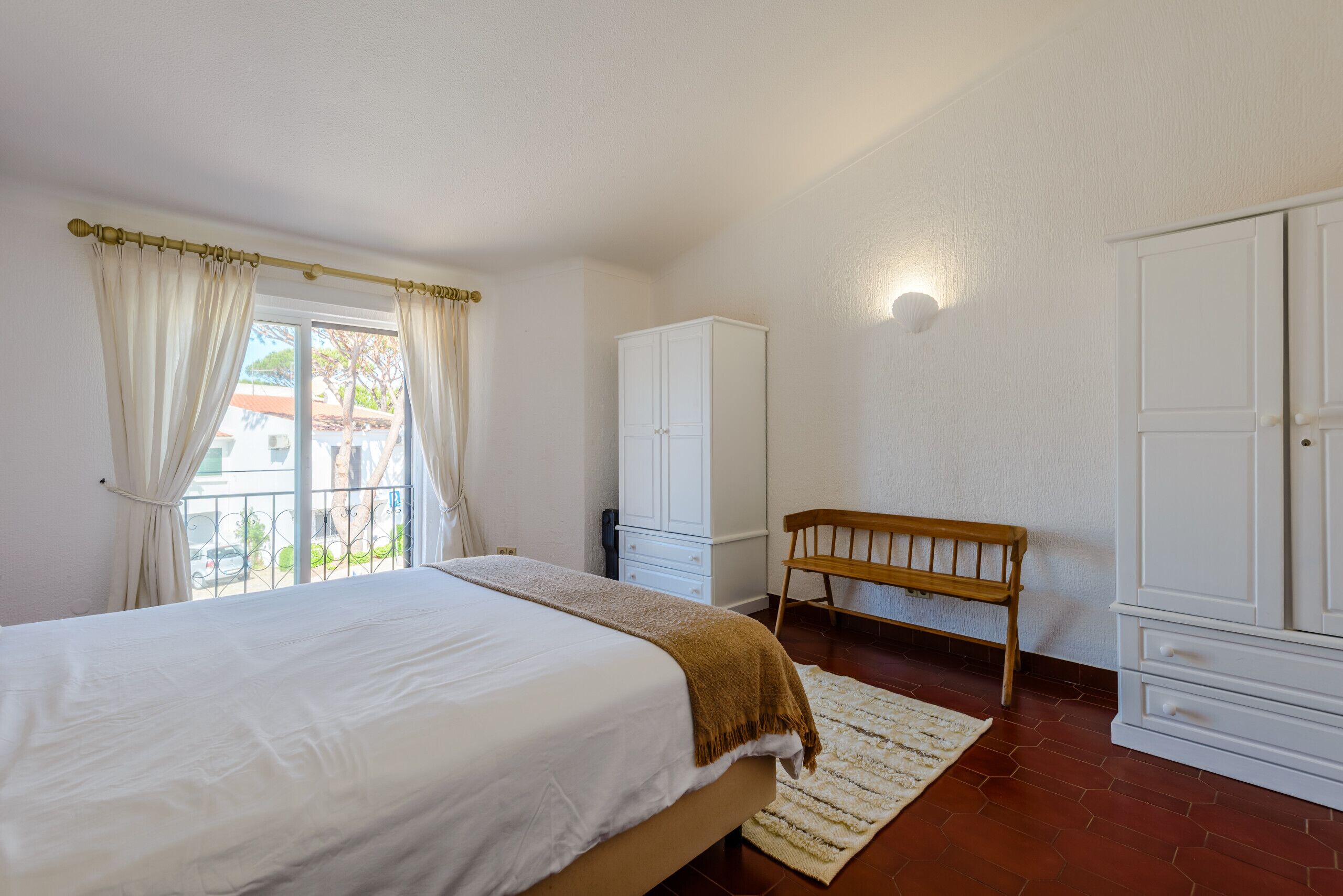 A bedroom with a bed, a wooden bench, a wardrobe, and a window with curtains.
