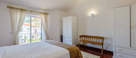 A bedroom with a bed, a wooden bench, a wardrobe, and a window with curtains.