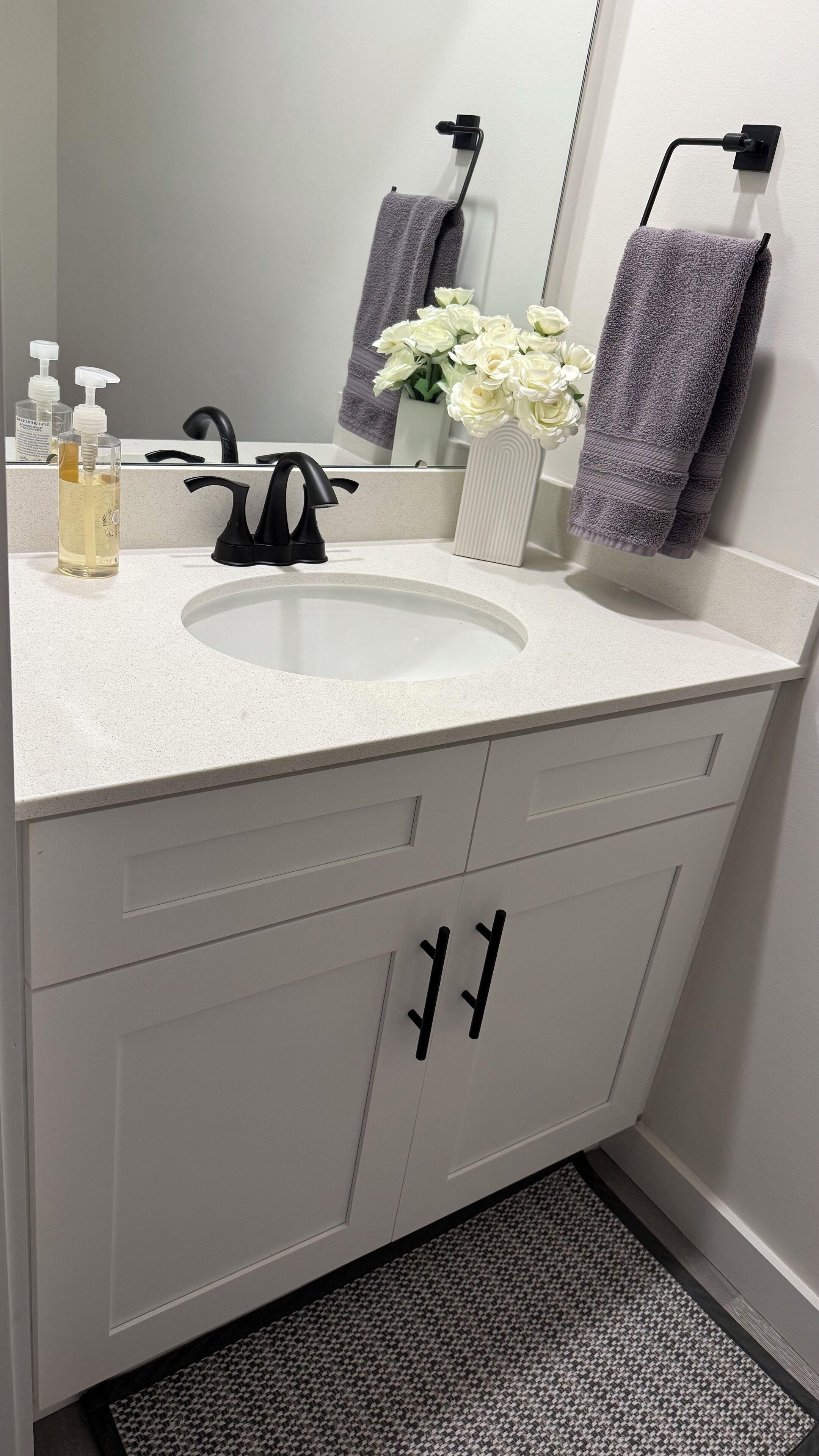 A bathroom with a white sink, black faucet, and two towels hanging on hooks. There is a bottle of hand soap and a vase with white flowers on the counter.
