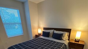 A bedroom with a bed, two bedside tables with lamps, a window with blinds, and a wooden floor.