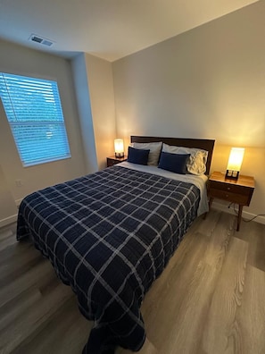 Une chambre à coucher avec un lit, deux tables de chevet avec des lampes, une fenêtre avec des stores et un plancher en bois.