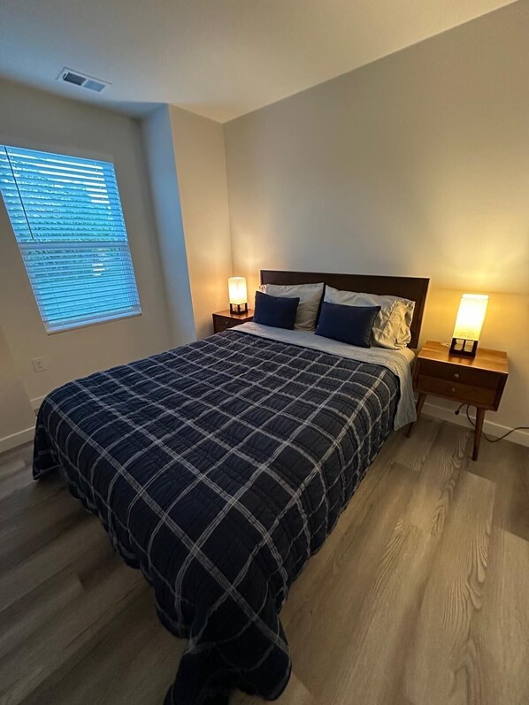 Quarto com cama, duas mesas de cabeceira com luminárias, janela com persianas e piso de madeira.