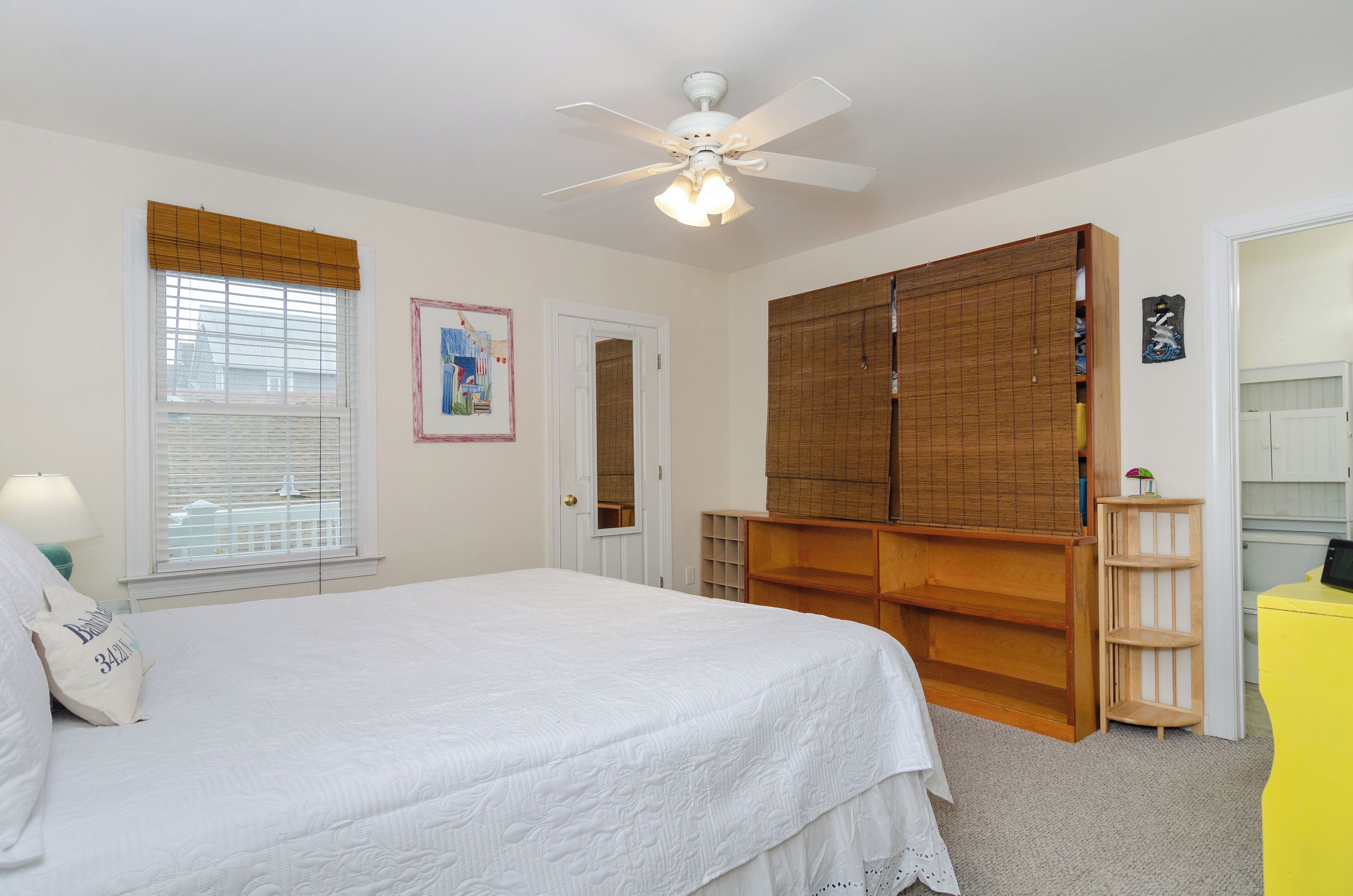 Une chambre à coucher avec un lit, un ventilateur de plafond, une armoire, une petite étagère et une porte.