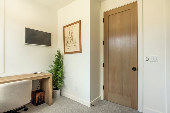 A room with a desk, a potted plant, a framed picture, and a wooden door.