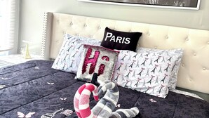 Ett sovrum med en säng som har ett täcke och kuddar med Paris-tema, en stor affisch med en stadsbild på väggen och en dekorativ utskärning av Eiffeltornet på sänggaveln.
