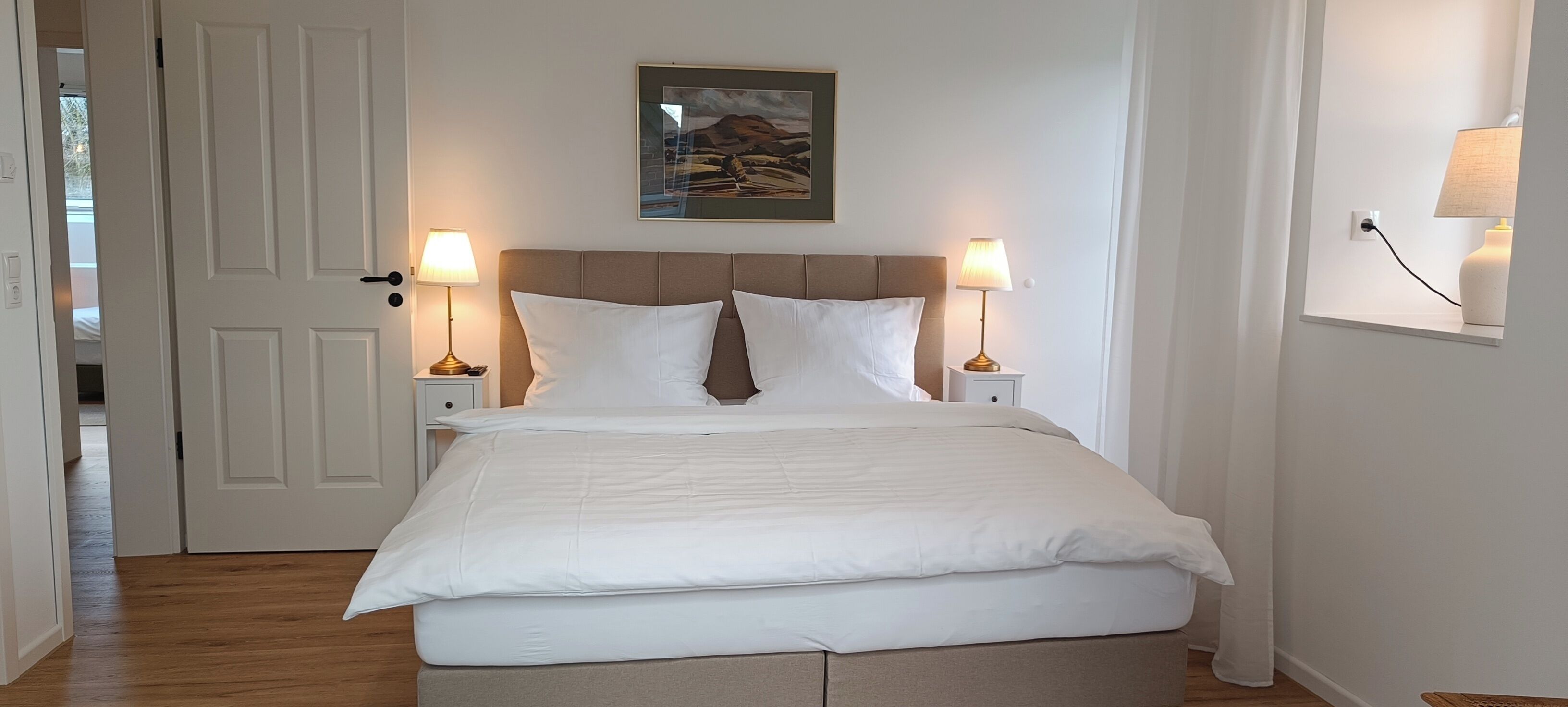 Una cama bien hecha con ropa de cama blanca, dos lámparas de noche, un cuadro en la pared y una puerta que da a otra habitación.