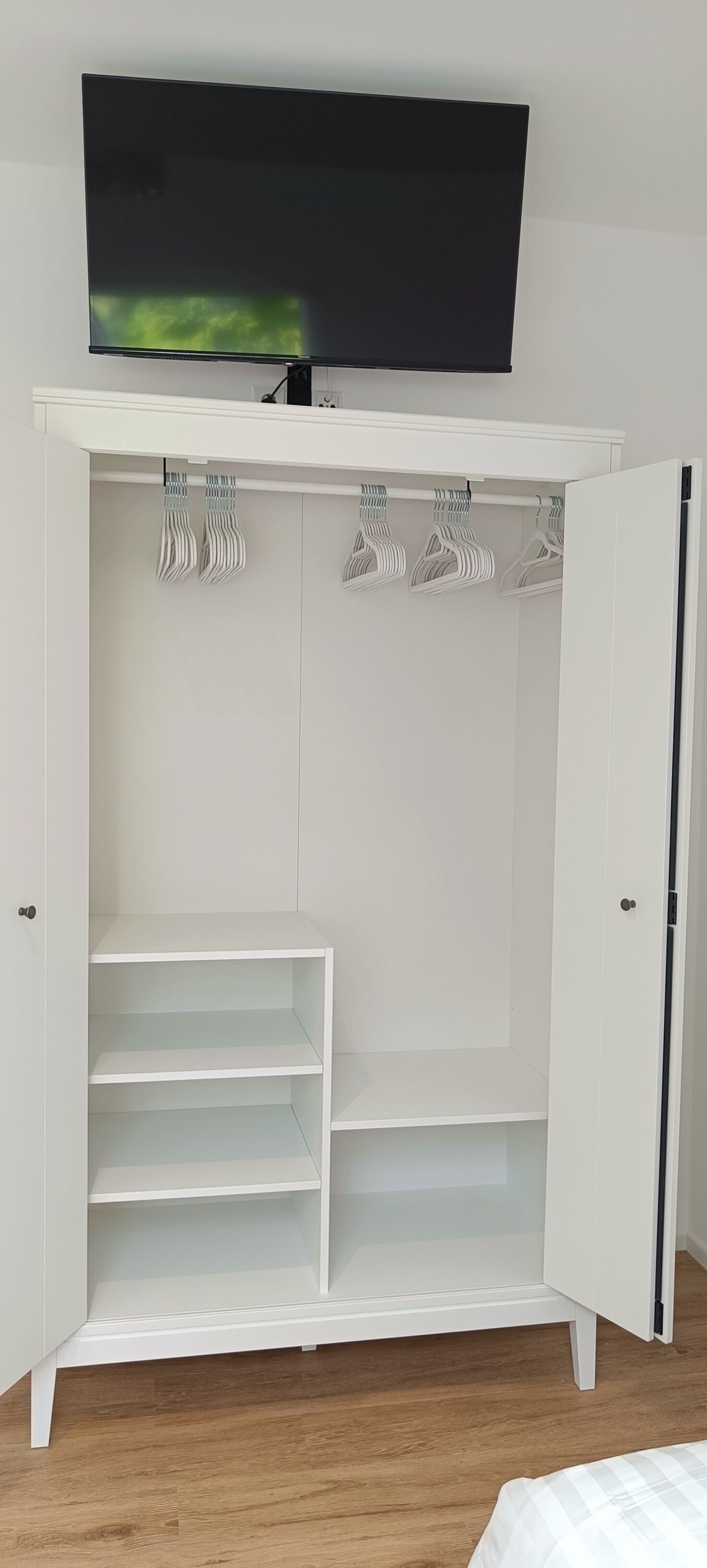 Un armario blanco con puertas abiertas, un televisor de pantalla plana montado arriba y una cama con ropa de cama blanca.