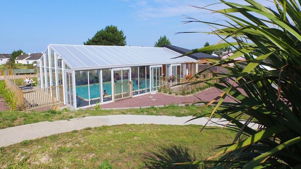 A glass-enclosed swimming pool with a transparent roof, surrounded by a wooden deck and a grassy area. - Charming Apartment for Five Guests (Port-Bail-sur-Mer)