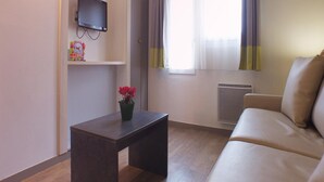 A living room with a sofa, a small table with a potted plant, a flat-screen TV mounted on the wall, and a window with curtains. - Charming Apartment for Five Guests (Port-Bail-sur-Mer)