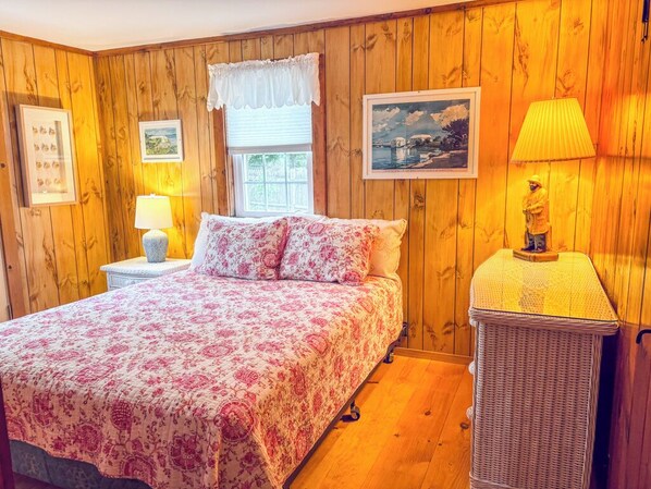 Une chambre avec un mur lambrissé en bois, un lit avec une literie à motifs floraux, une table de chevet avec une lampe et un fauteuil en osier.