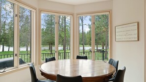 Un comedor con una mesa redonda de madera y sillas negras, ventanas amplias con vistas a árboles y una valla, y un letrero en el pared.