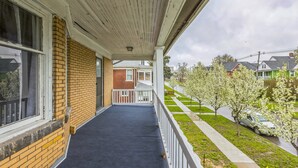 A covered porch with a brick wall, a black floor, and a view of a green house and trees in the background. - Comfy 3br Flat Close To The Falls 3 Bedroom Apts (Niagara Falls)