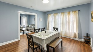 A dining area with a table set for six, wooden chairs, a white tablecloth, and a window with sheer curtains. - Comfy 3br Flat Close To The Falls 3 Bedroom Apts (Niagara Falls)