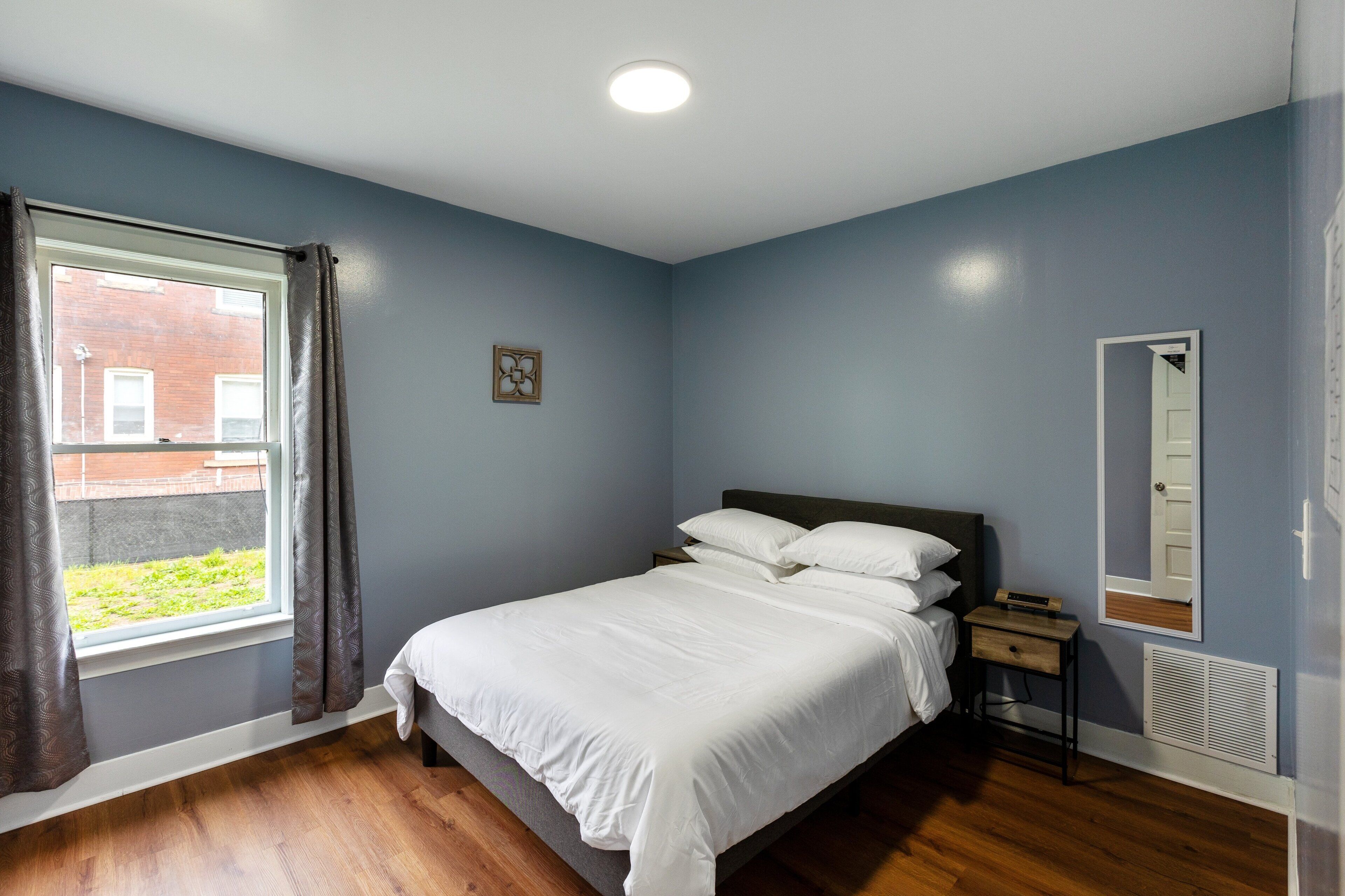 Une chambre à coucher comprenant un lit, une table de chevet, une fenêtre avec des rideaux, un miroir fixé au mur et un radiateur.