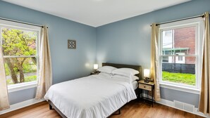 A bedroom with a bed, two bedside tables with lamps, a window with curtains, and a view of a brick building and greenery outside. - Comfy 3br Flat Close To The Falls 3 Bedroom Apts (Niagara Falls)