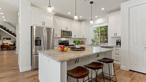 Une cuisine moderne avec des meubles blancs, des appareils en acier inoxydable, un grand îlot central doté de tabourets, un éclairage suspendu et une vue sur l’extérieur.
